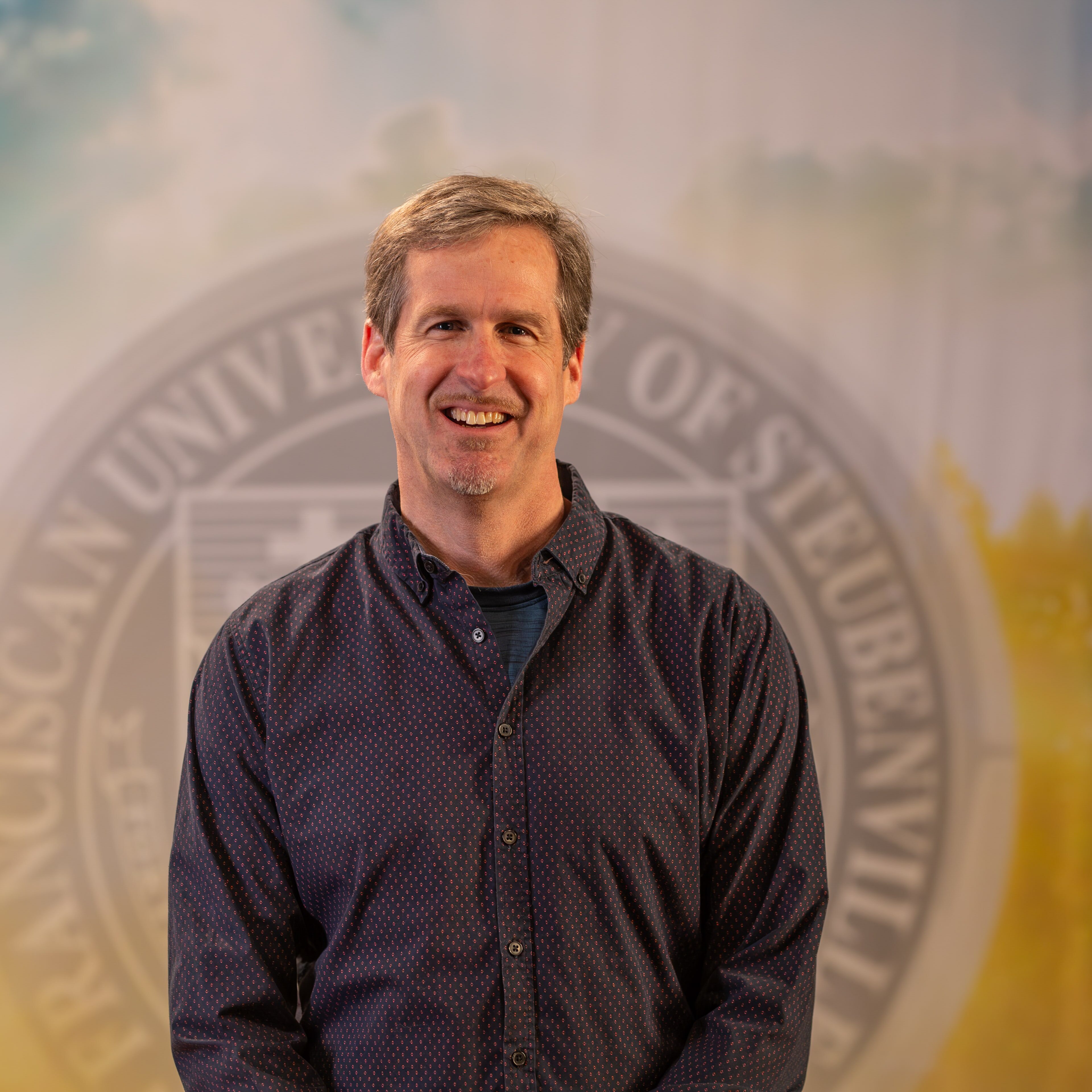 Dr. Dan Kuebler standing in front of the Franciscan University of Steubenville seal.