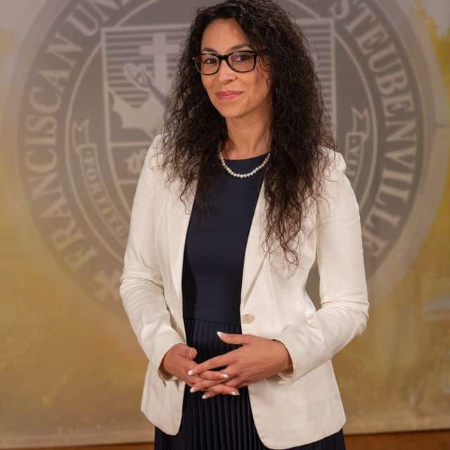 Dr. Fernanda Psihas wearing a dress and white blazer in front of the university seal.