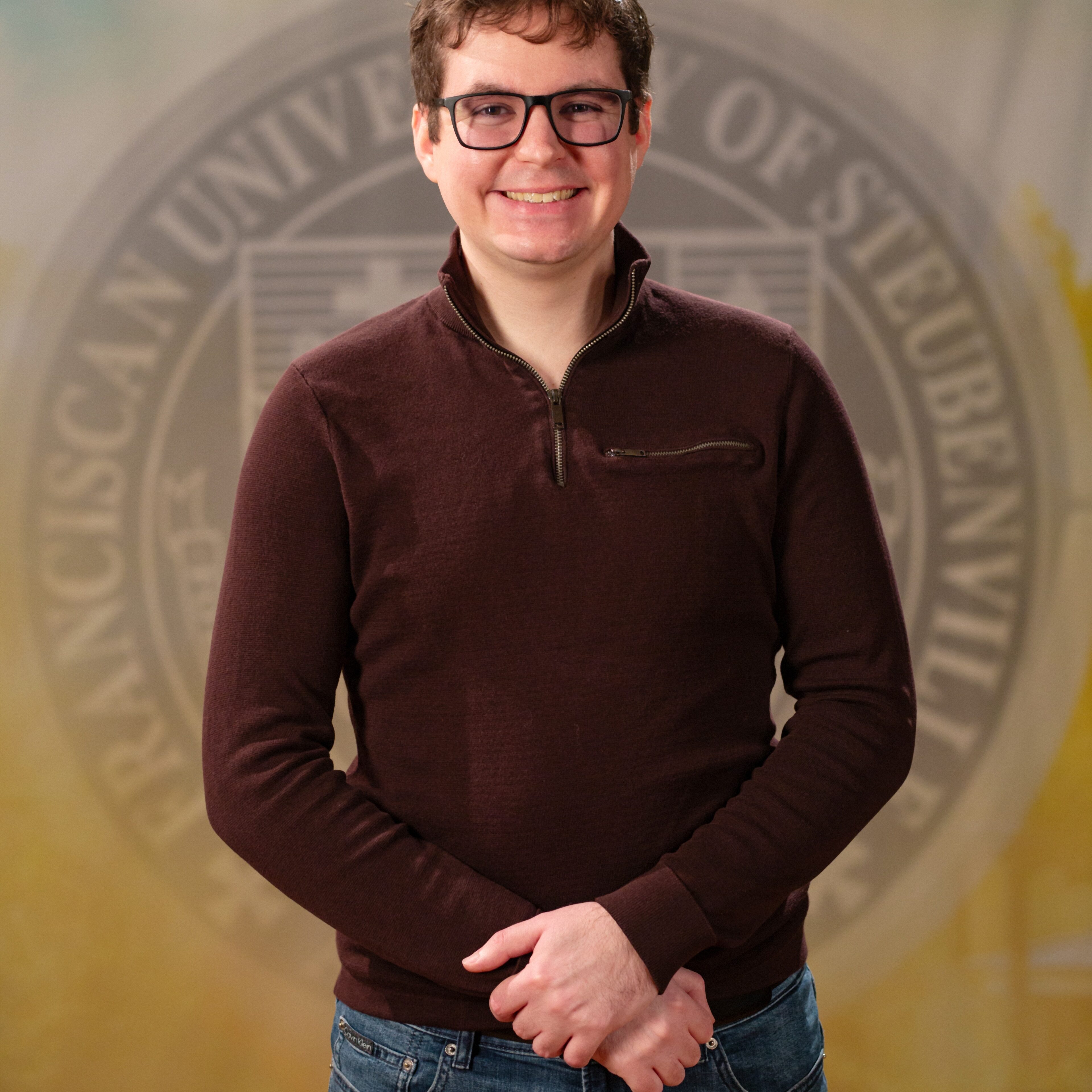 Faculty portrait of a Dr. Patrick Carzon standing in front of the Franciscan University of Steubenville seal.