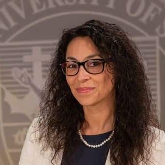 Dr. Fernanda Psihas wearing a dress and white blazer in front of the university seal.