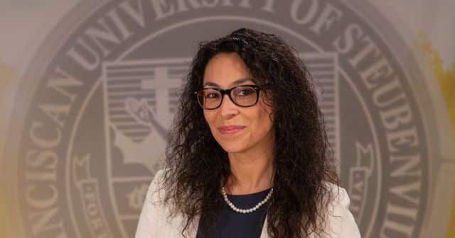 cropped-Dr-Fernanda-Psihas-2025.jpeg Dr. Fernanda Psihas wearing a dress and white blazer in front of the university seal.